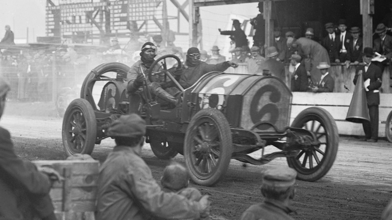 The Simplex 90 races at the Vanderbilt Cup in 1910, black and white photo with two dirt-covered men behind the wheel as the car speeds past the finish line