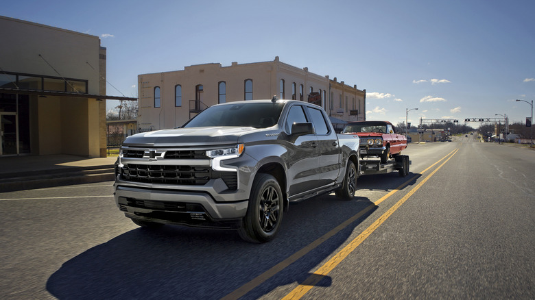 Gray 2026 Chevrolet Silverado 1500 pickup truck seen in left front three-quarter view towing a vintage car on a trailer