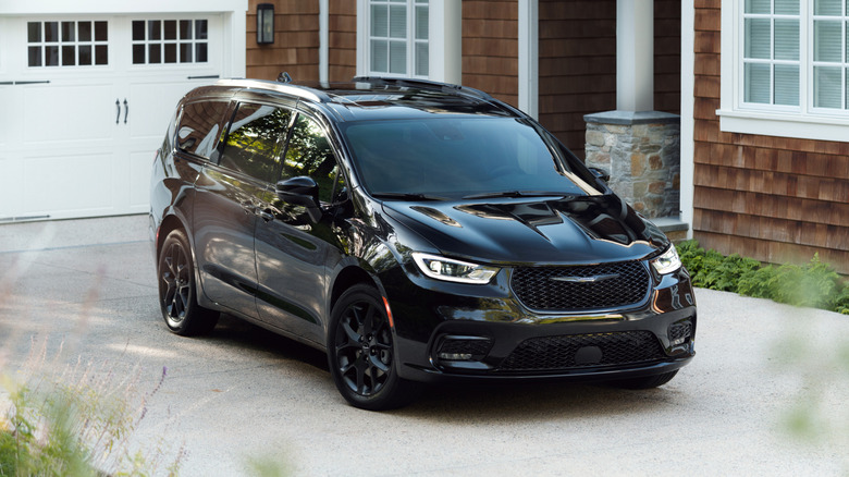Black 2026 Chrysler Pacifica minivan seen in right front three-quarter view parked in the driveway of a wood-shingle house