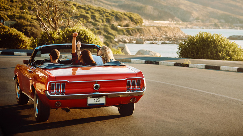 Stock image of three people in a classic Ford Mustang Convertible driving near water.