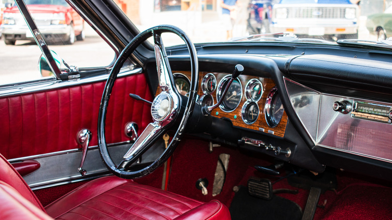 The interior of a classic car, featuring chrome dials in a wooden dashboard, a bright black steering wheels, and red leather seats.