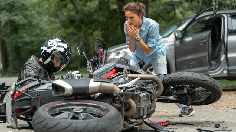 Aftermath of crash involving motorcycle. Concerned female driver stands over motorcyclist who is on the ground.