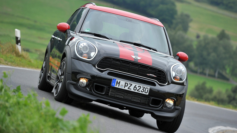 A front shot of a black and red 2012 Mini Countryman JCW driving around a corner on a rural road surrounded by grass and trees