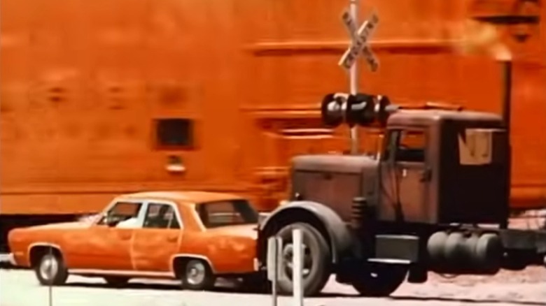 A screenshot from the official movie trailer for the 1971 film "Duel" showing a Peterbilt semi-truck pushing a Plymouth Valiant toward a moving train.