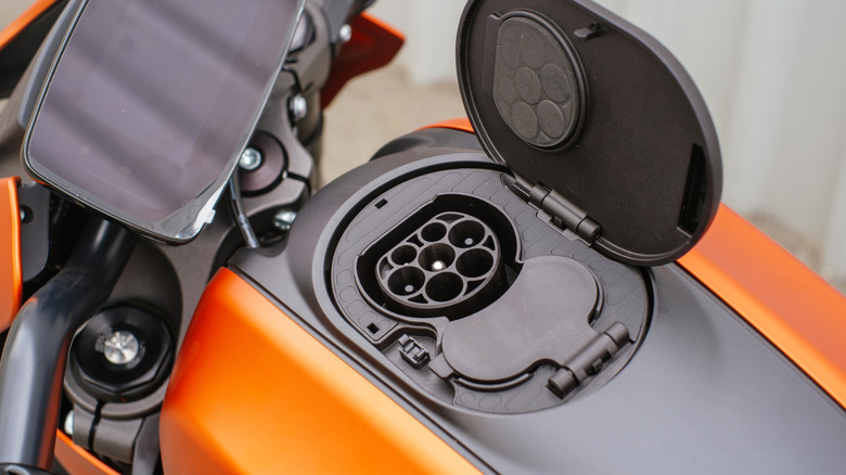 An electric motorcycle's charging port is shown close up with the door open against orange bodywork.