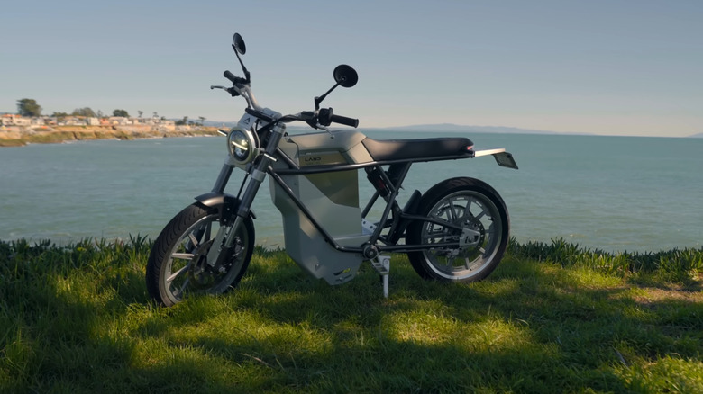 LAND District electric motorcycle sits in grass near a body of water in the shade.