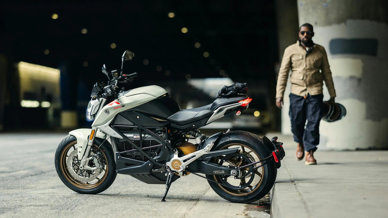 An electric motorcycle sits near a curb as a man with a helmet approaches from behind.