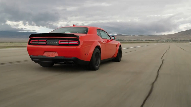 A reddish Dodge Challenger speeds down a runway with mountains far ahead.