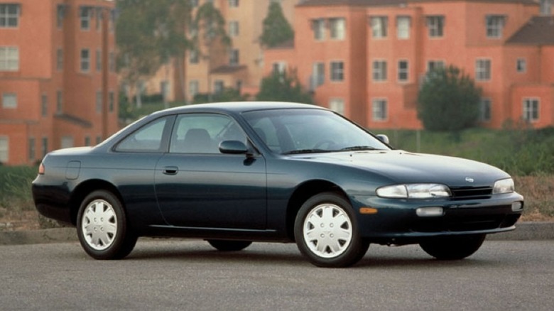 A dark green 1996 Nissan 240SX coupe parked in front of terracotta residential buildings.
