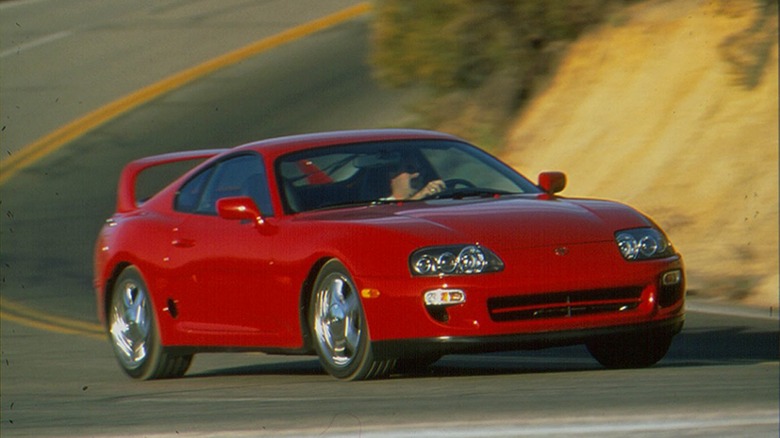 A red 1998 Toyota Supra Turbo with a high rear wing driving along a winding mountain pass road.