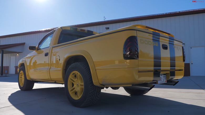 A yellow Dodge Dakota R/T pickup truck with black racing stripes is shot from the rear in an industrial parking lot.