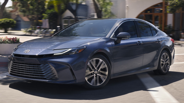 A blue 2026 Toyota Camry in a parking lot in front of trees and a building.