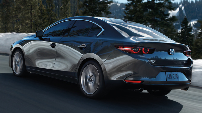 A gray Mazda Mazda3 Sedan in motion on a roadway while passing trees and snow-covered ground.