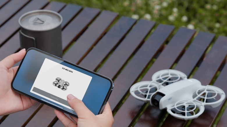 A person controlling a DJI Neo using a smartphone app, while the drone and a can sit nearby on a slotted table
