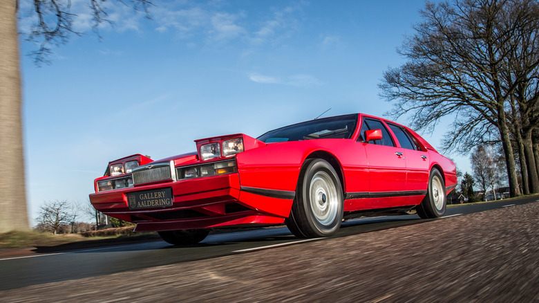 A low-angle dynamic shot of a red Aston Martin Lagonda driving on a country road