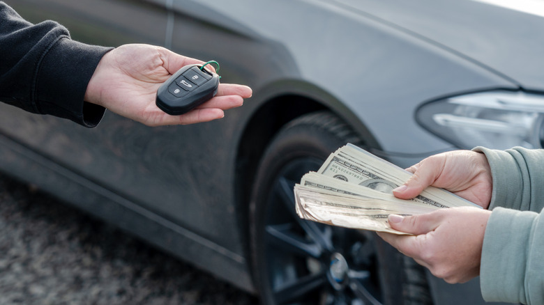 A buyer and a seller exchange cash for keys in front of a grey sedan.