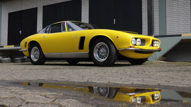 A shot of a yellow Iso Grifo taken from near the ground, with the car's reflection showing in a puddle in the foreground and a brick building in the background