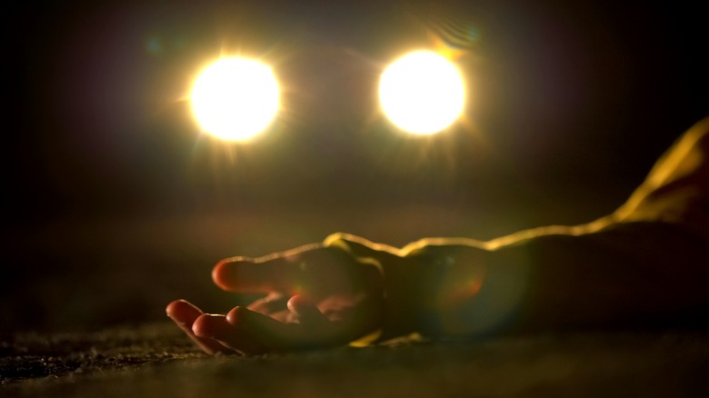 A hand lying on the road before a car's headlights