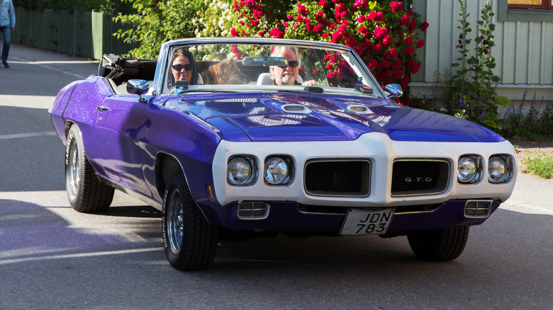 A vibrant purple-colored standard model 1970 Pontiac GTO being driven to a car show.