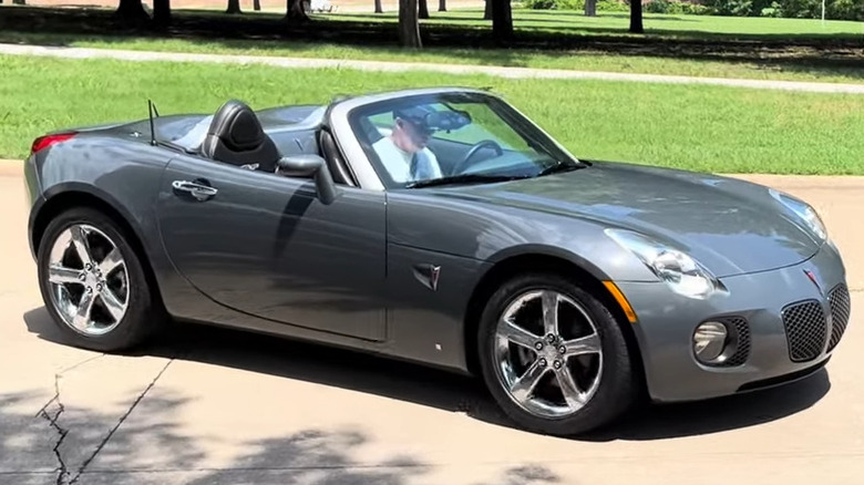 A gray Pontiac Solstice GXP roadster driving with the top down on a paved path.