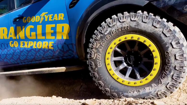 A Goodyear Wrangler Boulder MT tire mounted on a wheel with a bright yellow beadlock ring kicking up dust on a trail