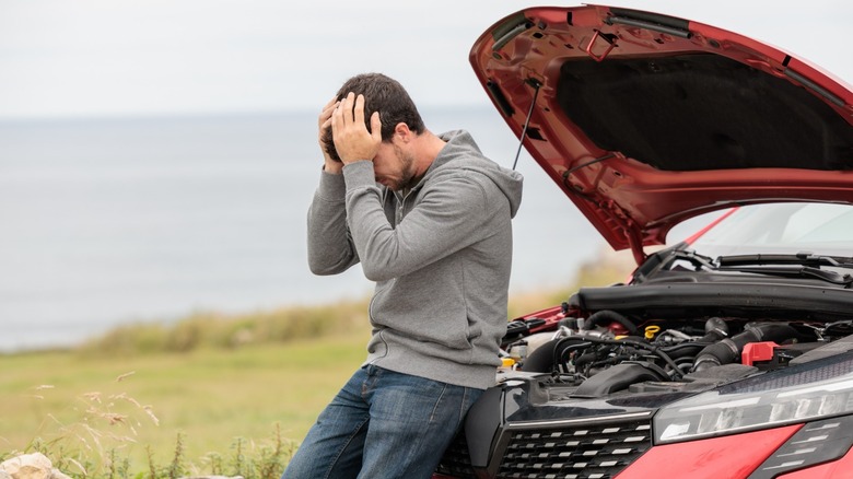 A man holds his head in his hands while leaning against a car that is broken down on the roadside