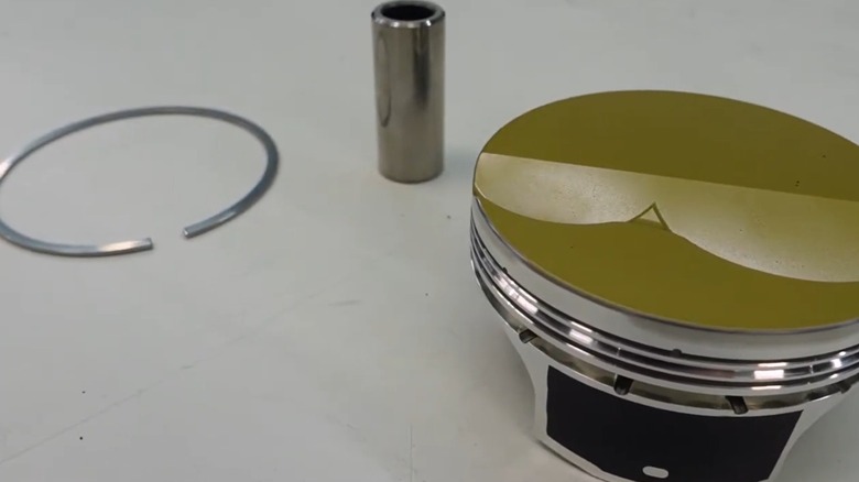 A piston ring, wrist pin, and piston on a flat white surface