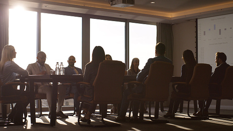 A shadowy boardroom discussion taking place near large windows with a graph projected on one wall