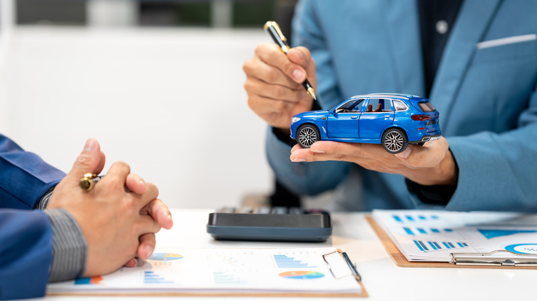 A man in a suit gesturing with a pen as he holds up a blue hatchback model at a table
