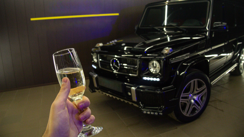 A person's hand holding up a glass of champagne in front of a black Mercedes G-Wagon