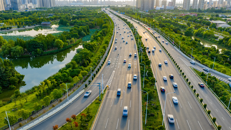 A busy highway with a variety of vehicles shown from above, with surrounding greenery and a skyline visible