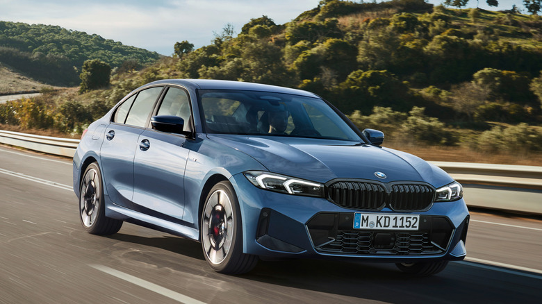 A blue BMW 330i on a curving roadway with tree-covered hills in the background