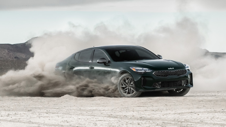 A dark-green 2023 Kia Stinger in motion and kicking up a cloud of dust on a gravel surface.