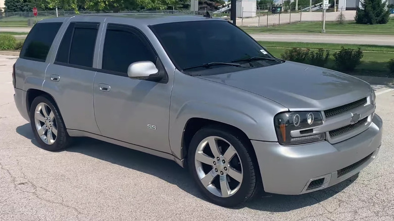 A Trailblazer SS in silver, parked on cracked asphalt near grass, trees and a building