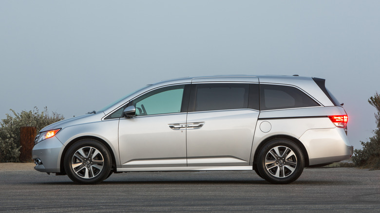 A silver 2015 Honda Odyssey minivan seen from directly left parked with shrubbery in background