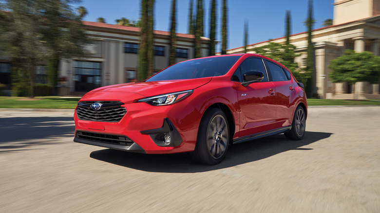 Red Subaru Impreza hatchback left front three-quarter view driving in front of collegiate-looking buildings