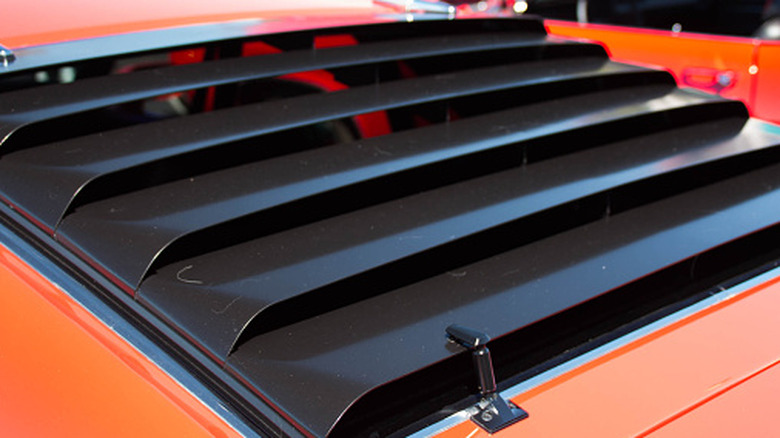 The rear window of an orange car is covered by black window louvers.