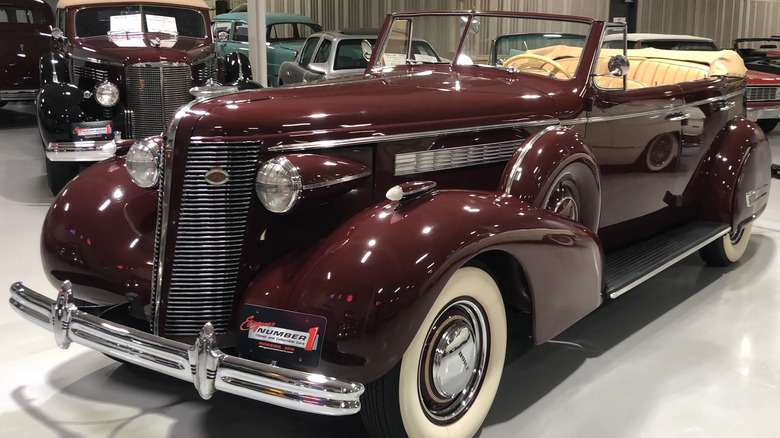 A maroon 1938 Buick Roadmaster 80-series "C body" convertible sedan standing amid other cars and wearing whitewall tires.