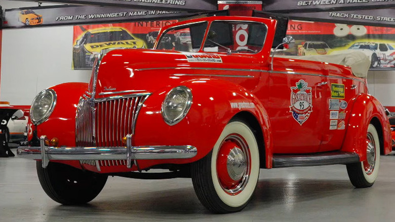 A bright red 1939 Ford De Luxe convertible sedan, standing amidst other cars indoors.