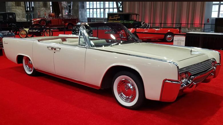 A cream-colored 1961 Lincoln Continental convertible standing inside a large building on red carpeting.