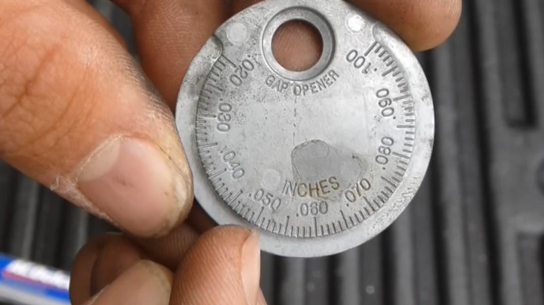 A close-up image of a person's hand holding a spark plug gap tool and its measurements.