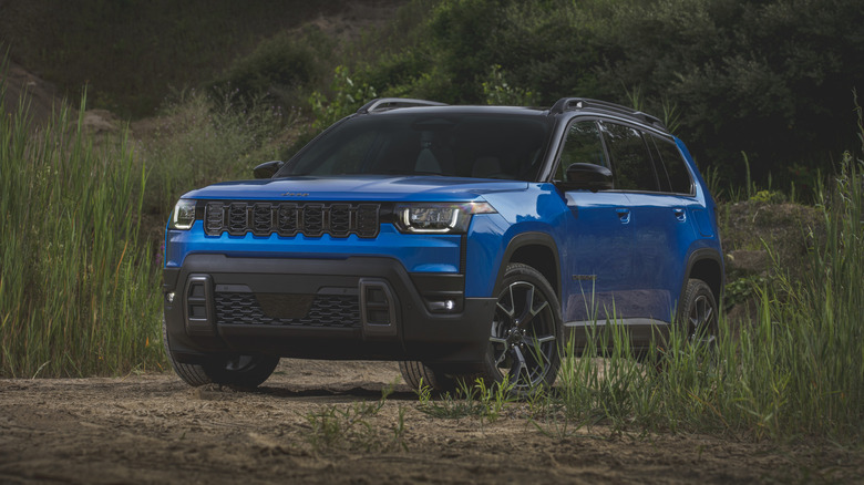 Blue 2026 Jeep Cherokee SUV left front three-quarter view seen on a dirt road  by tall grass