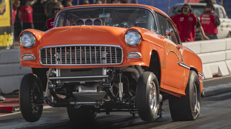 Orange Chevy gasser doing a wheelie during a drag race, with pit crew workers in the background behind a safety barrier.