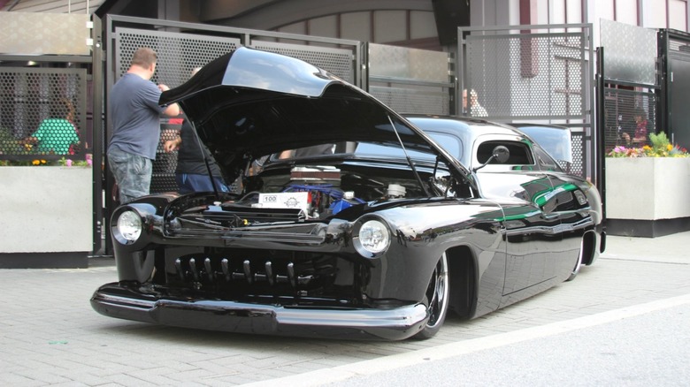 A black-colored lead sled hot rod with an open hood on display outdoors with people standing around the vehicle.