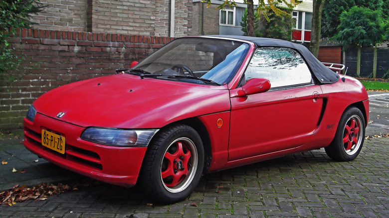 A red 1992 Honda Beat stis parked halfway on the sidewalk on a city street.