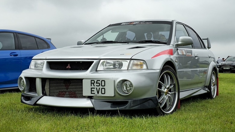 A silver 1999 Mitsubishi EVO VI sits on grass under cloudy skies.