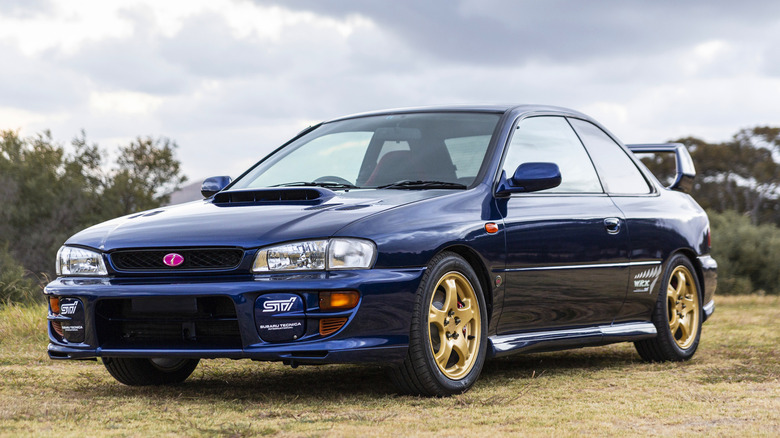 A blue WRX STI coupe with gold wheels sits parked on grass.
