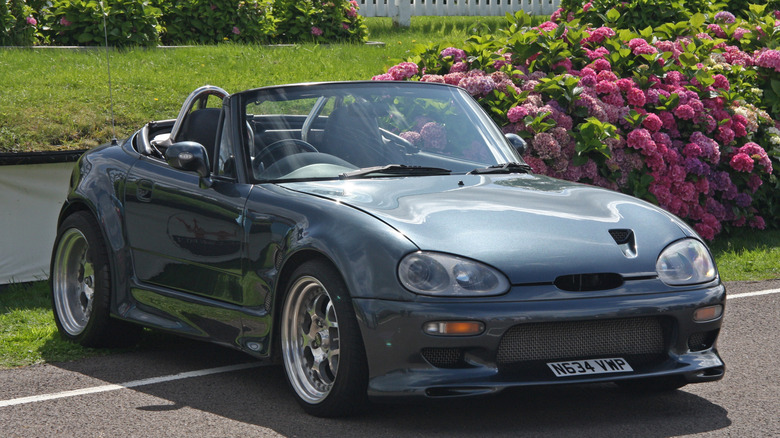 A light blue Suzuki Cappuccino sits parked near flowers.
