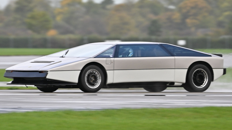 A wedge-shaped ultra-low 1979 Aston Martin Bulldog concept car, finished in its restored two-tone silver and dark grey paint scheme, speeding down a damp airfield runway.