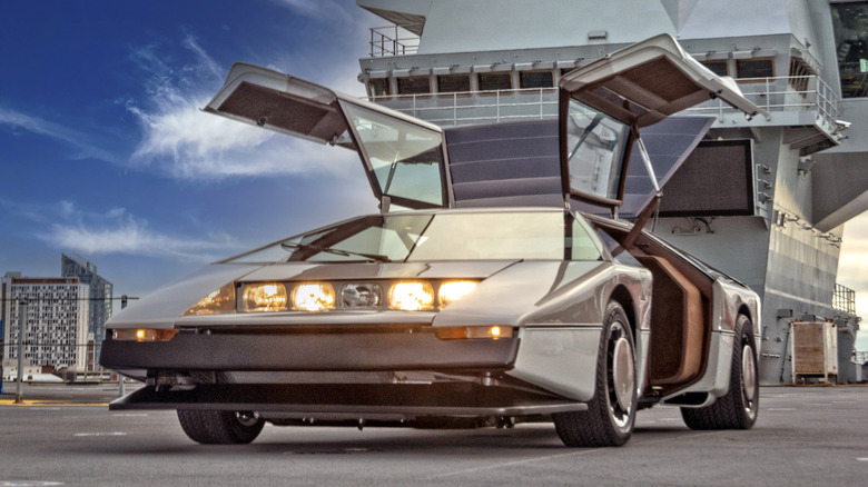 A metallic silver 1979 Aston Martin Bulldog concept car parked on the deck of an aircraft carrier with its gullwing doors fully open and its five center-mounted headlights illuminated.